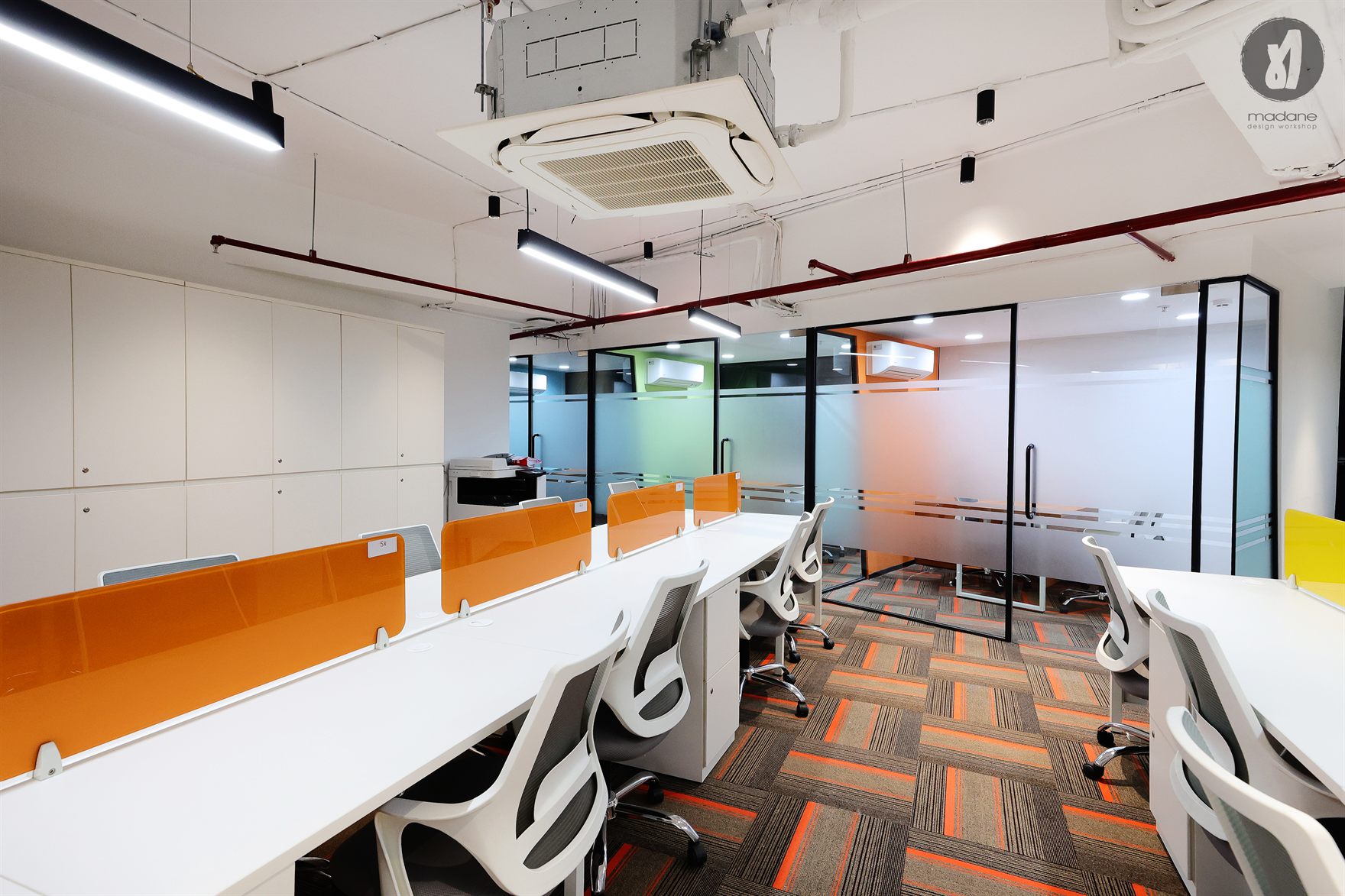 Open floor of white desks with orange screens beside colour-blocked glass cabins on orange-striped carpet under exposed ceiling