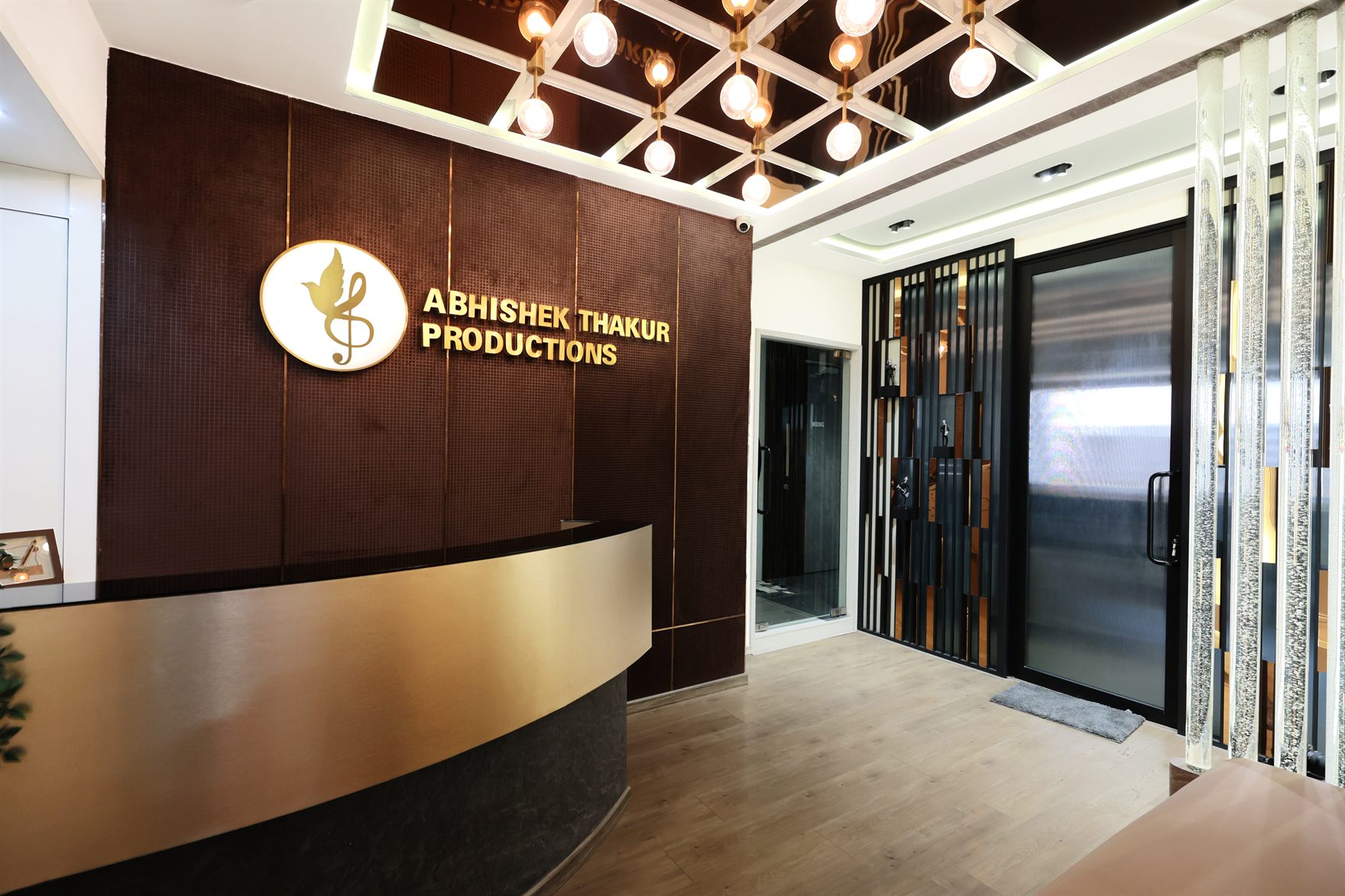 Studio reception with a curved brass-and-black stone desk against a dark textured feature wall carrying a gold treble-clef bird logo, under a mirror-bronze coffered ceiling set with globe filament pendants