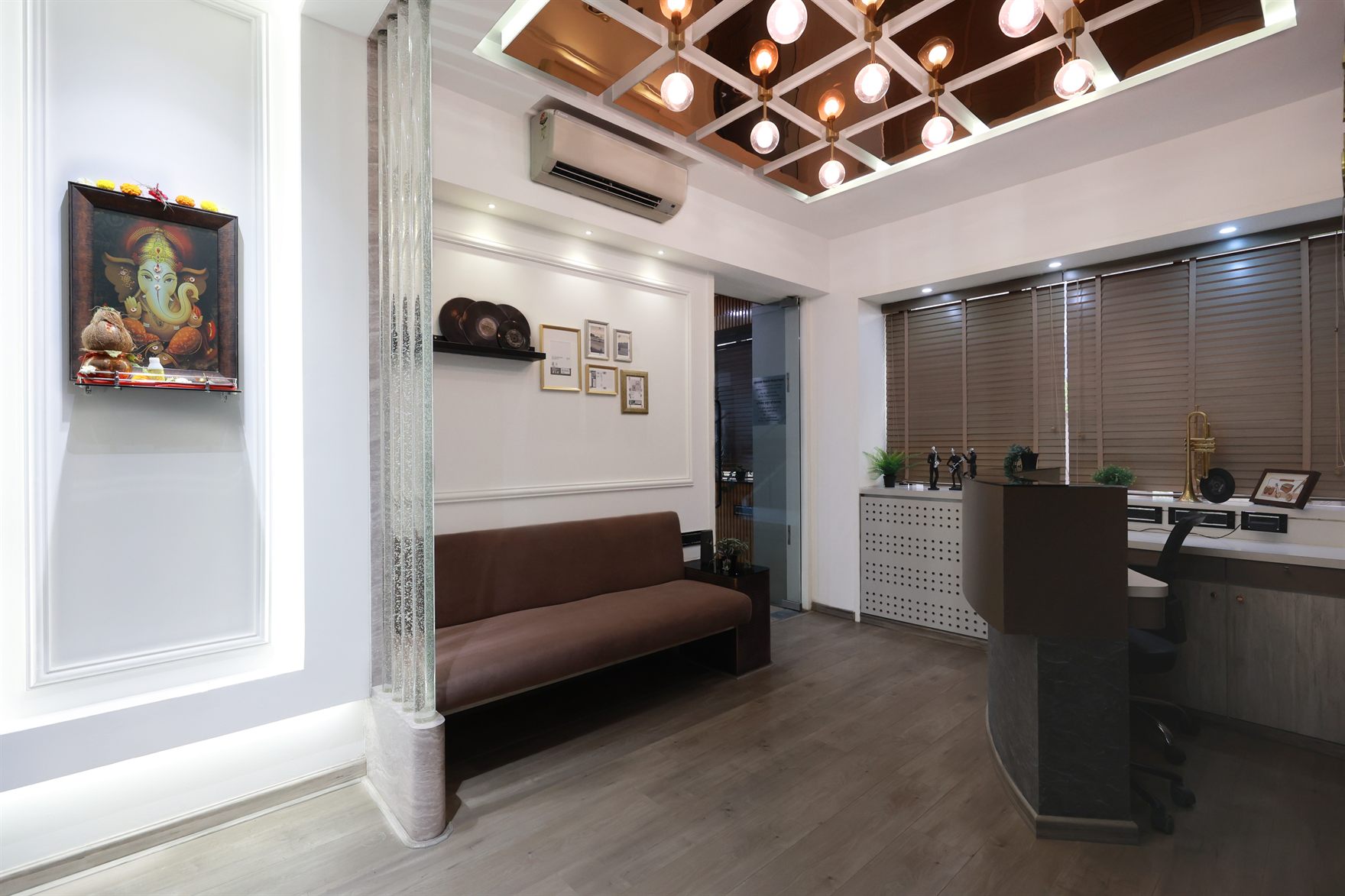 Waiting lounge with a brown sofa below a framed Ganesha, timber venetian blinds and a bronze-mirror coffered ceiling of globe lights beside a compact reception desk