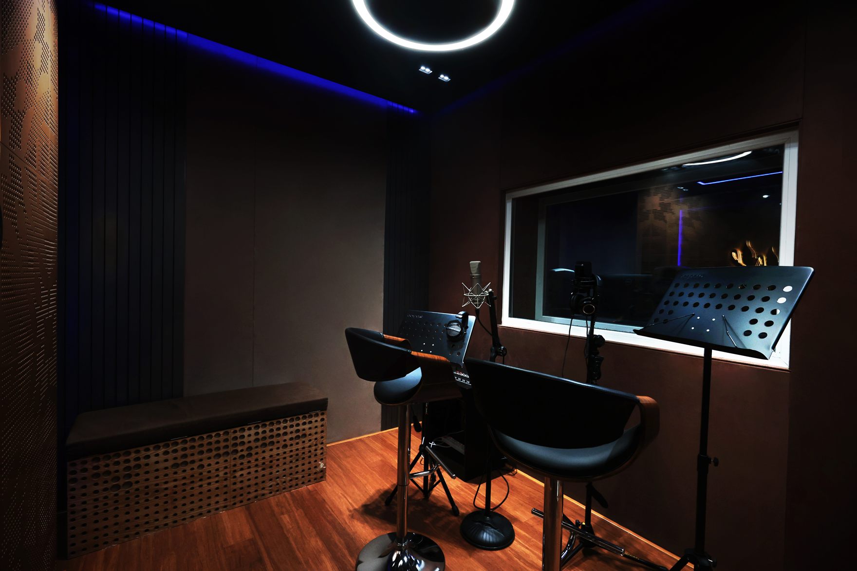 Vocal recording booth with dark acoustic slat and perforated-copper panels, a circular LED ring light, two black stools and mic and music stands facing a window into the control room