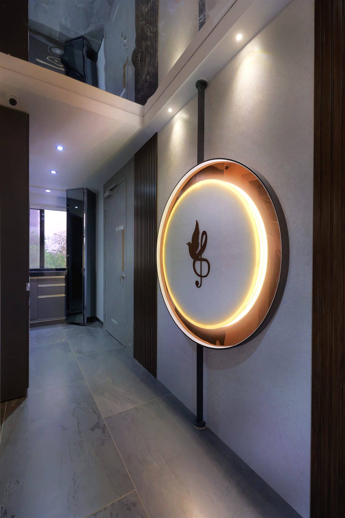 Double-height corridor with a backlit circular sign bearing a bronze bird-and-treble-clef emblem on a grey plaster wall, fluted timber panels and marble-look tile flooring