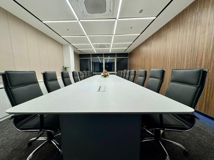 Boardroom with long white conference table, executive black leather chairs, warm wood-panelled wall and grid-recessed linear lighting.