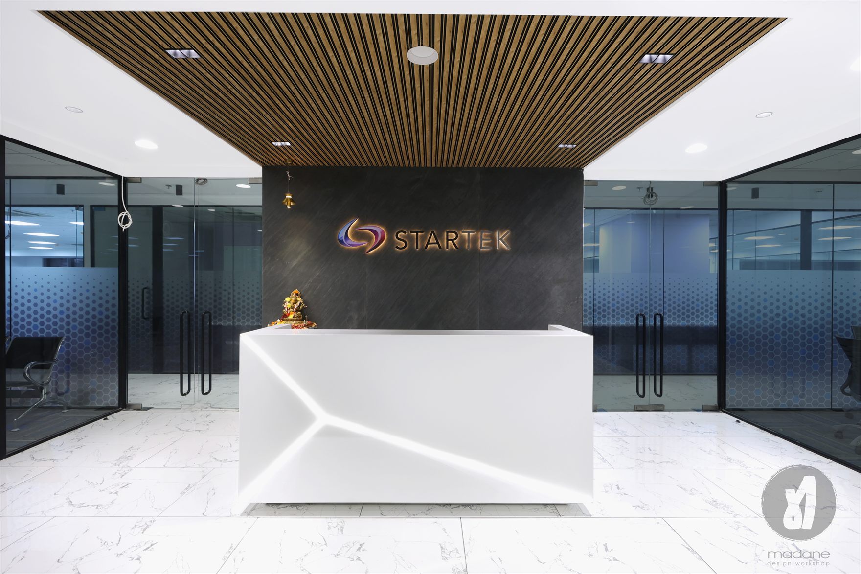Startek reception with a backlit STARTEK logo on a dark stone wall, a sculptural white desk under a timber-slat ceiling and glazed offices with dotted film on either side