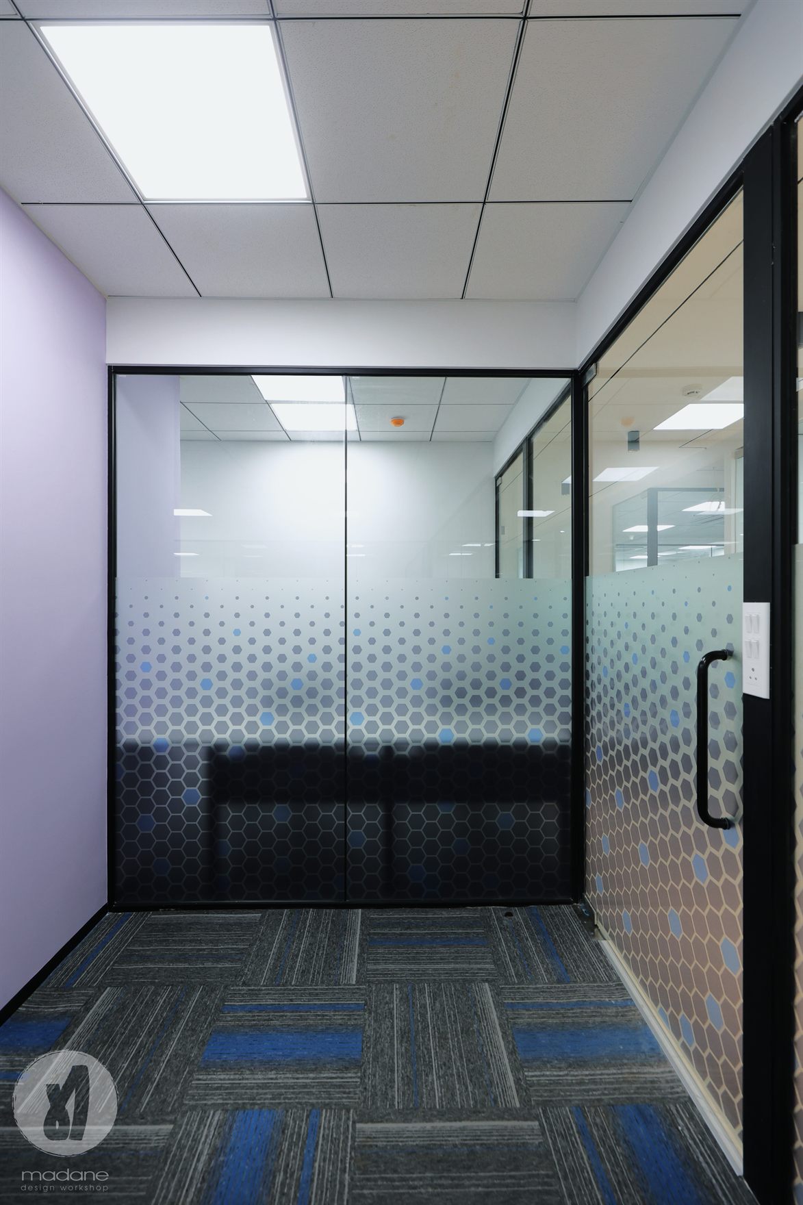 Compact glass-walled cabin with lilac wall and blue hexagon film partition on blue-striped carpet tiles