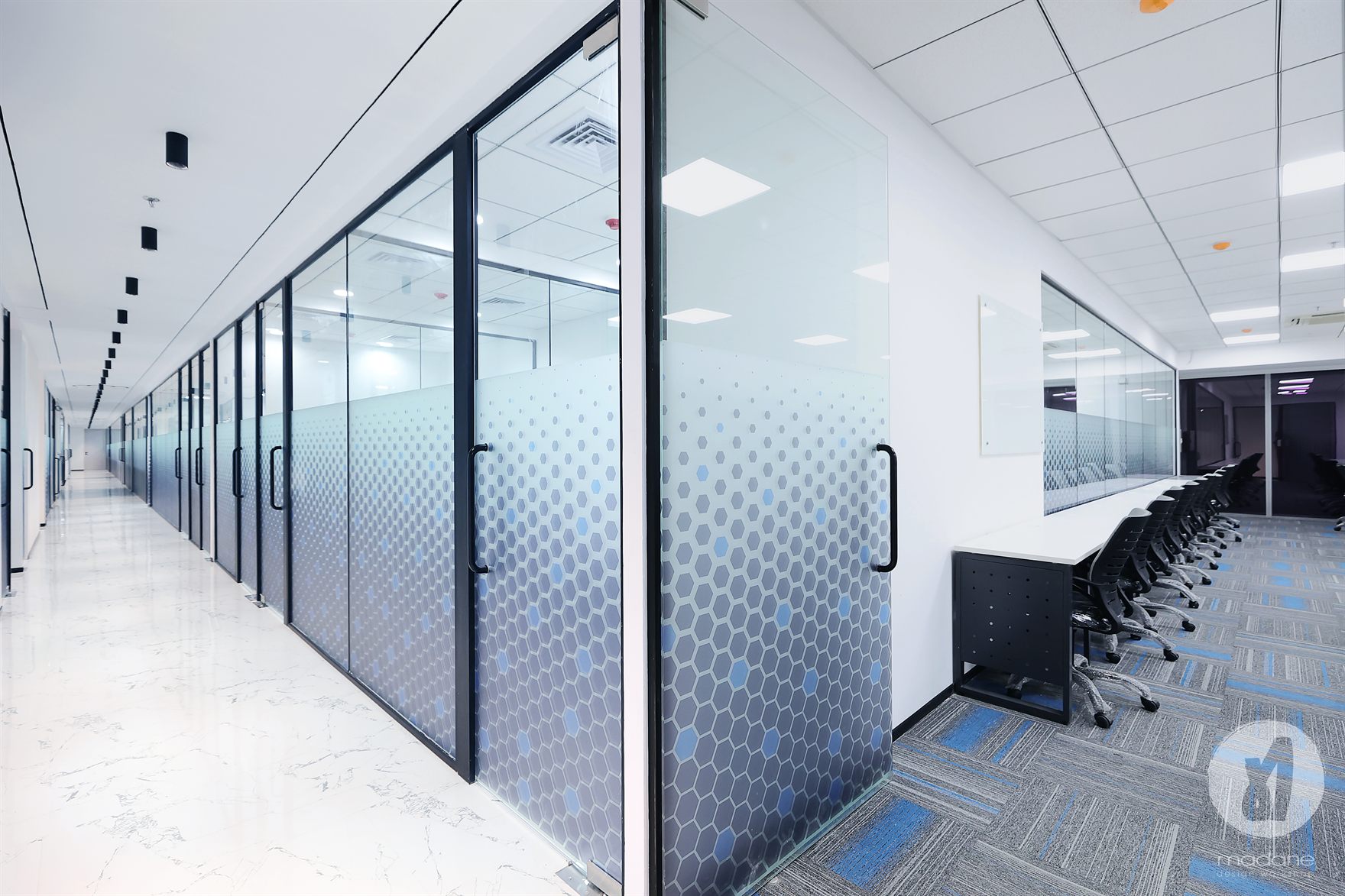 Corridor between marble flooring and a bench of black chairs, flanked by glass cabins in blue hexagon frost film