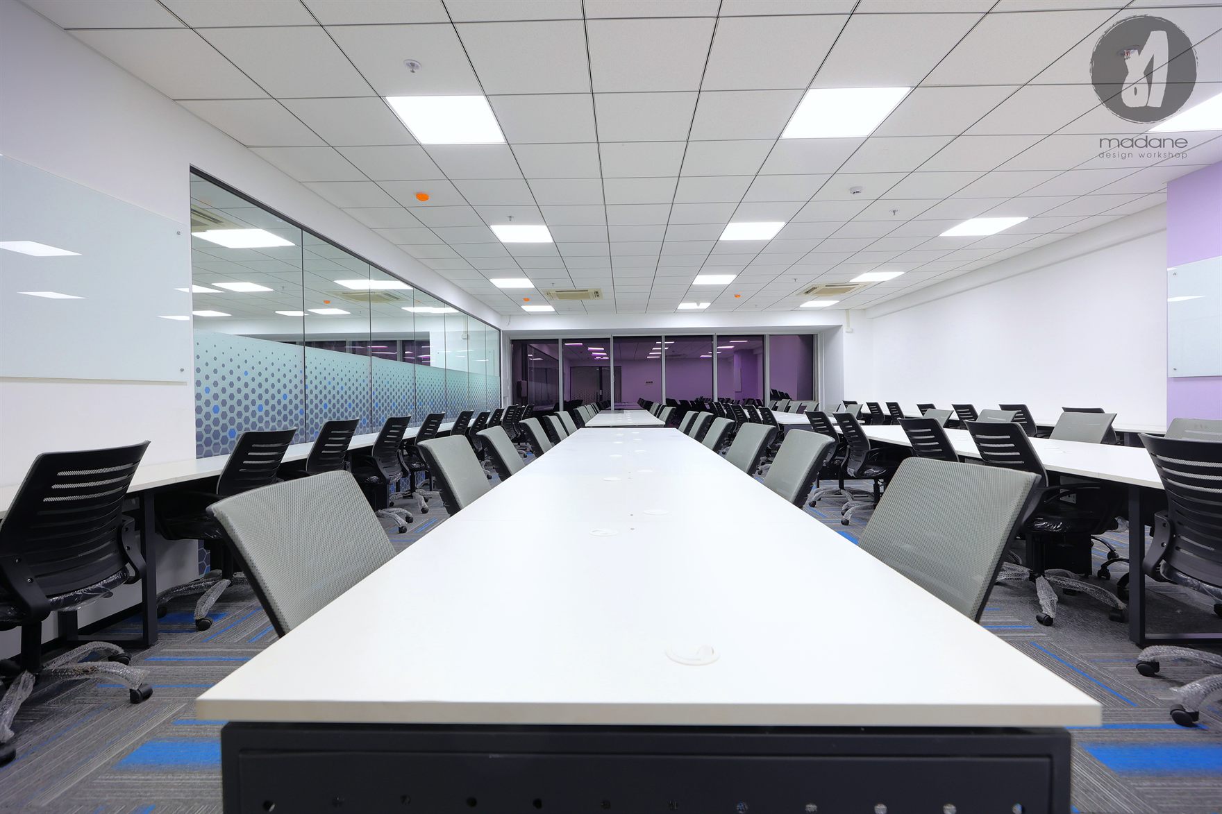 Long white training tables lined with grey and black mesh chairs, glass hexagon-film partition on one side and a purple wall beyond
