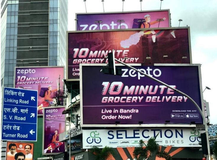 Cluster of purple Zepto billboards advertising 10 minute grocery delivery on a Mumbai street above a Selection Bikes storefront