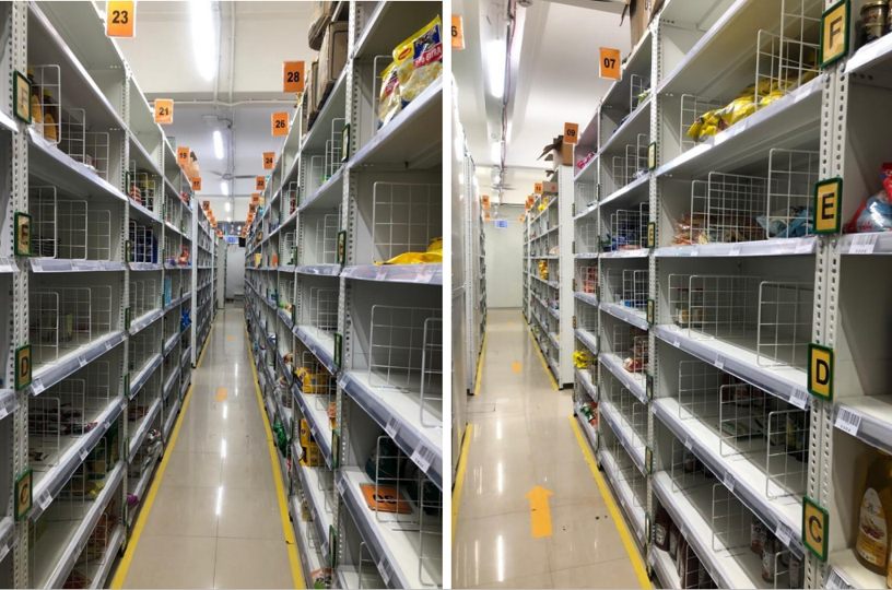 Picking aisle detail of colour-coded wire-mesh shelving holding grocery stock, with lettered shelf zones and directional yellow floor arrows on a tiled floor